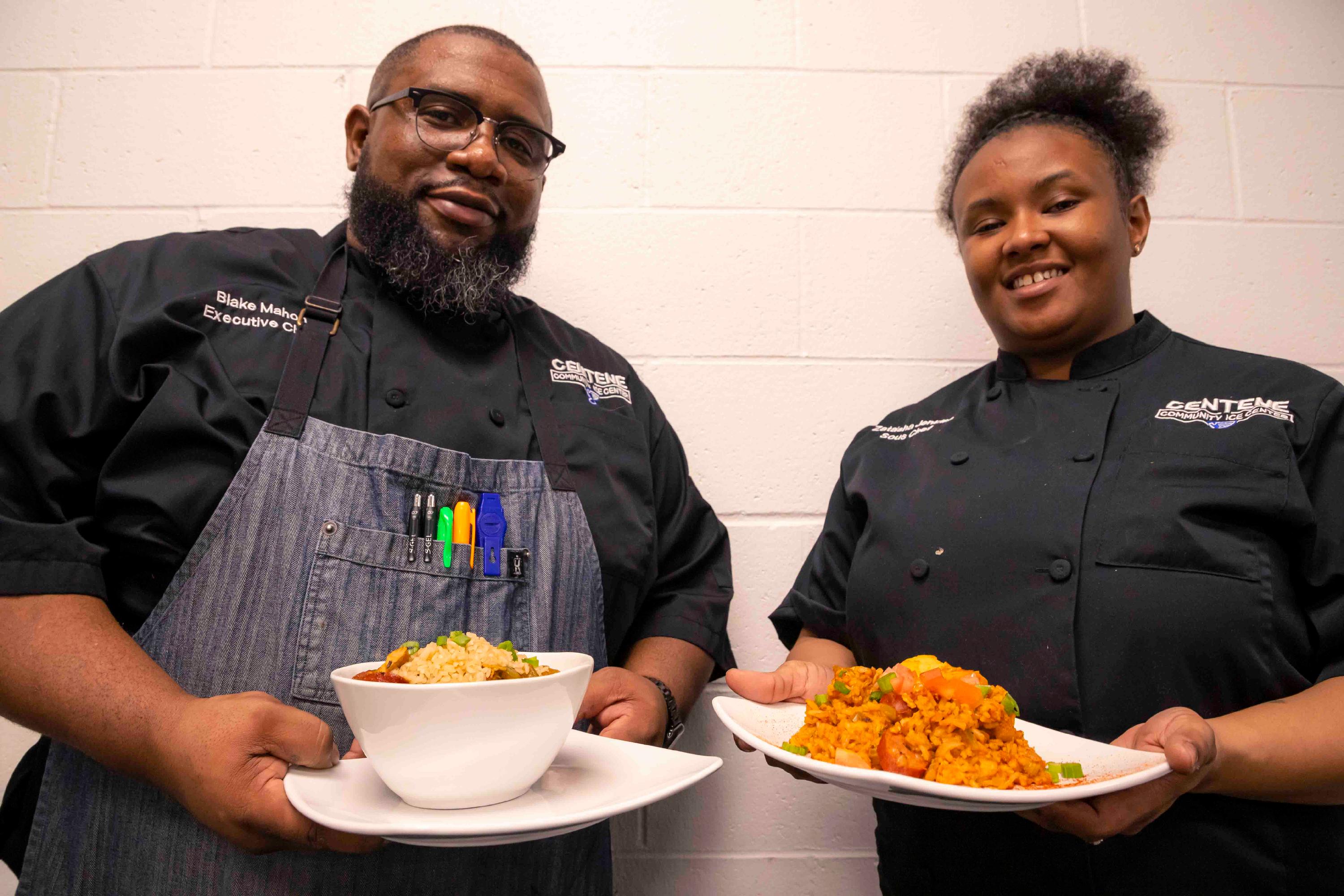 A male and female chef wearing Centene Community Ice Center Uniforms hold a plate of gumbo and jambalaya, respectively, while smiling down at the camera